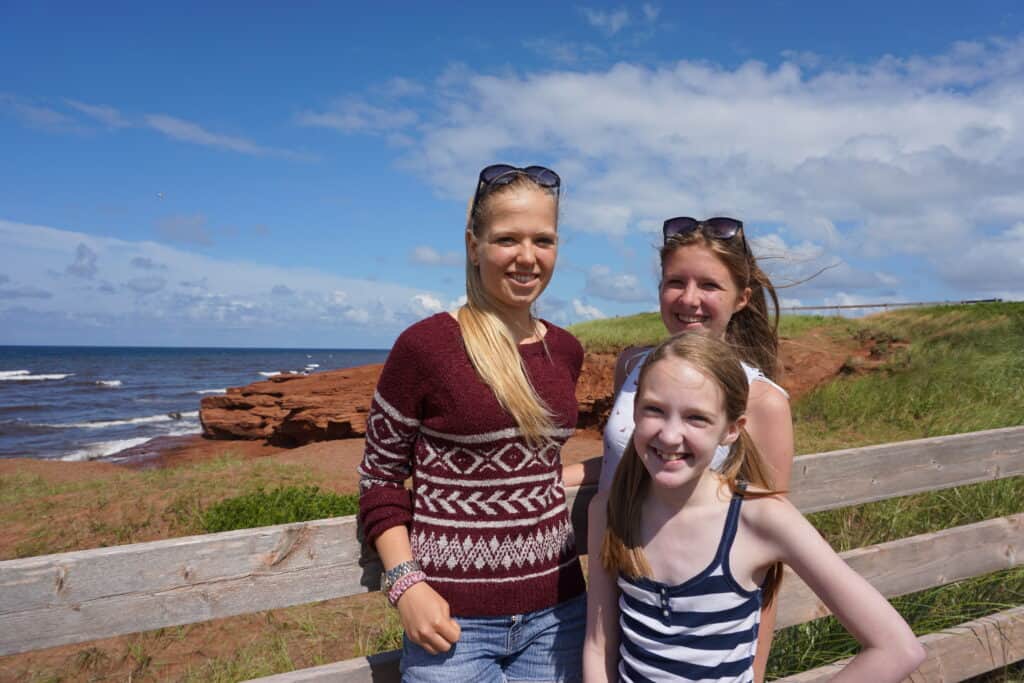Two teenage girls and a younger girl standing in front of wooden fence with sand dunes, red cliffs and ocean in background - Prince Edward Island National Park.