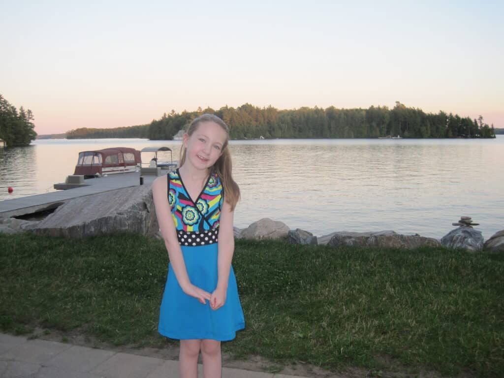 young girl in colourful dress stands in front of Lake Rosseau with dock and small boat in background at dusk.