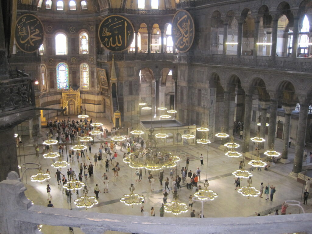 Looking down from the second level of Hagia Sophia at main level with hanging chandeliers and groups of people.