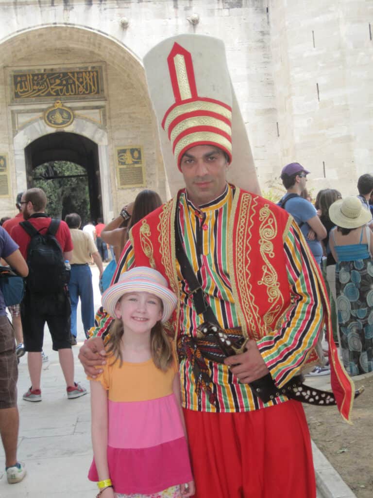 In midst of crowd at Topkapi Palace, young girl poses with man dressed as Sultan - colourful red, gold and striped costume and red and gold striped headpiece.