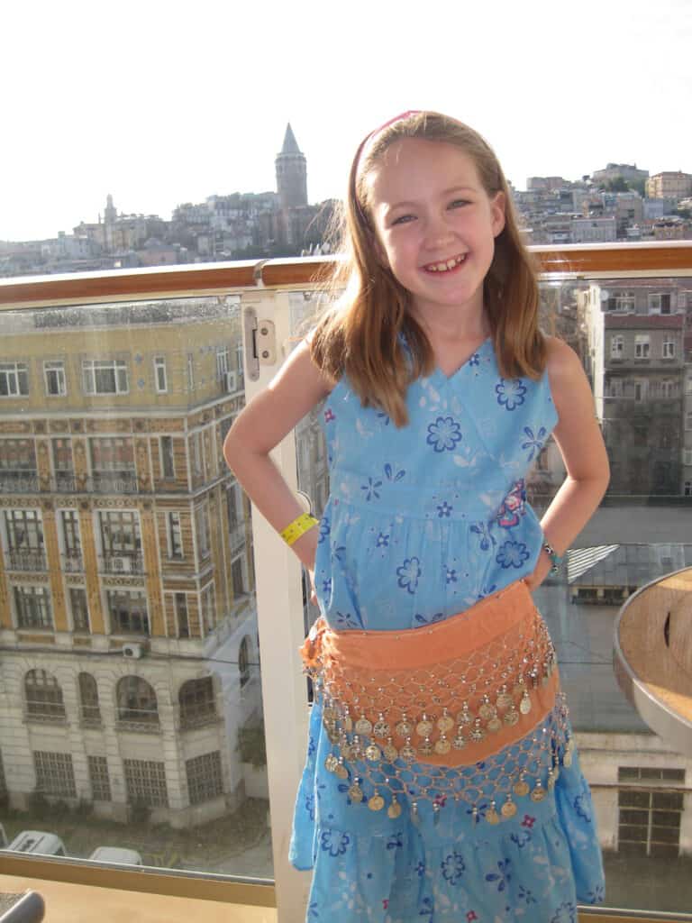 Young girl in blue dress wearing orange scarf with metal rings around her waist posing on cruise ship balcony with buildings and Galata Tower in background.