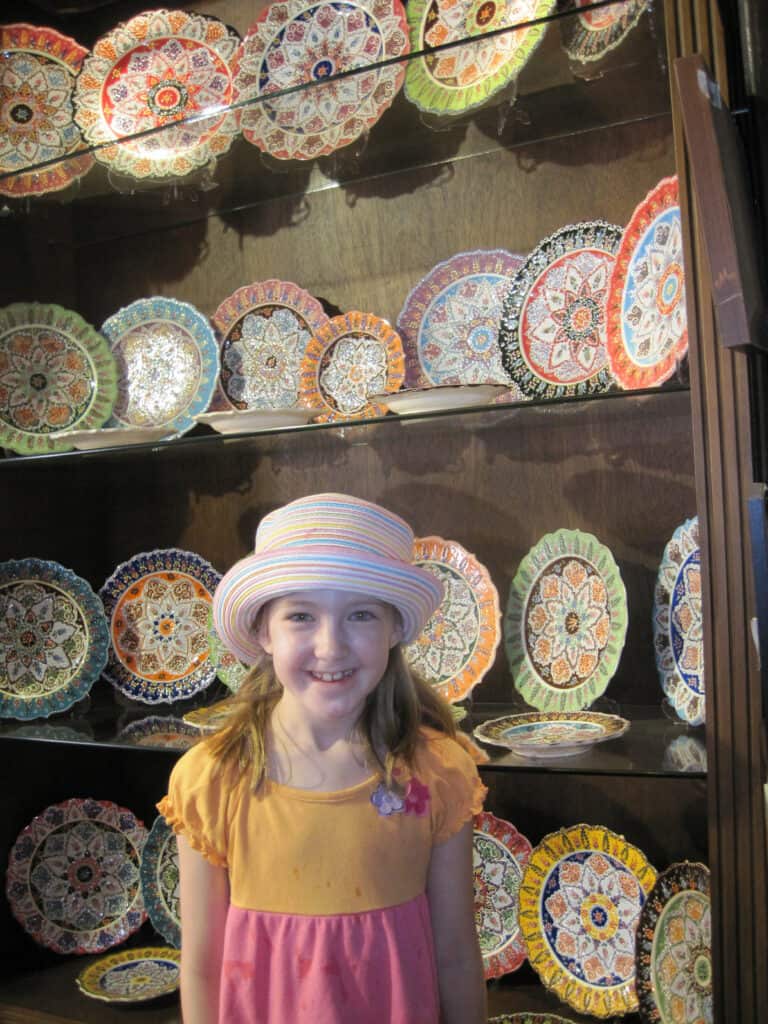 Young girl in pink and orange striped top and striped multicoloured hat posing in front of cabinet of bright and colourful ceramic plates in Istanbul.