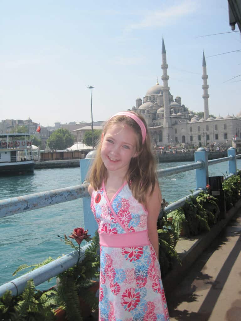 Young girl wearing pink and blue flowered dress and pink headband posing along the Bosphorus with buildings in background.
