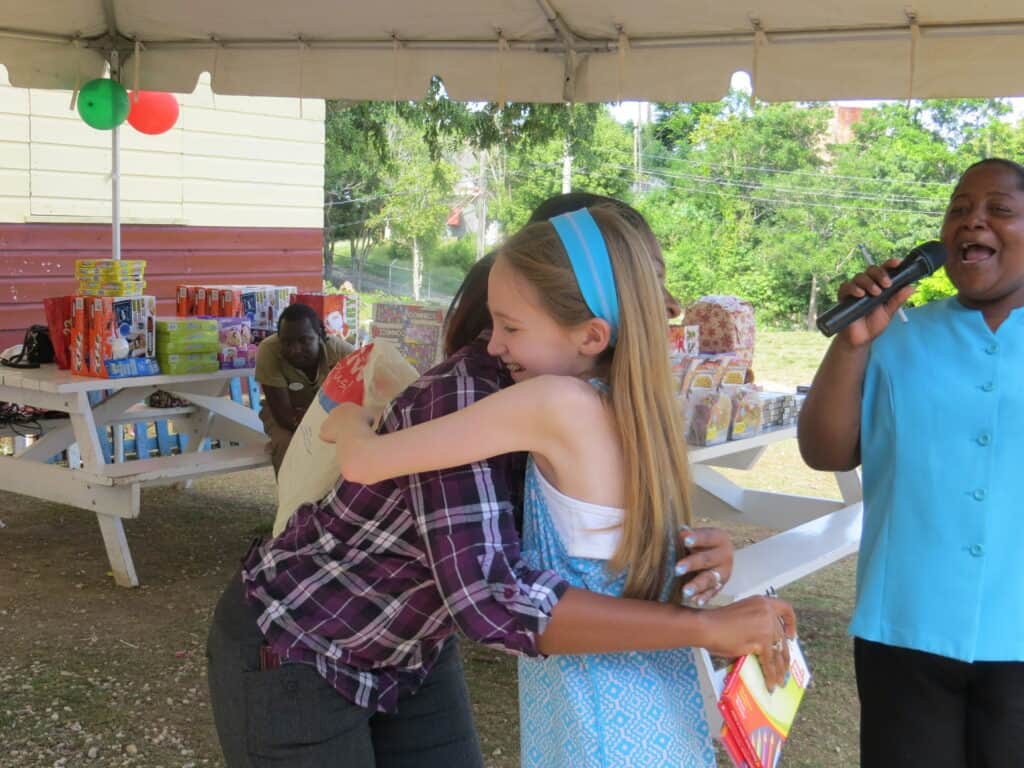 girl in blue and white dress hugs woman in plaid shirt while in background woman in blue shirt holds microphone.