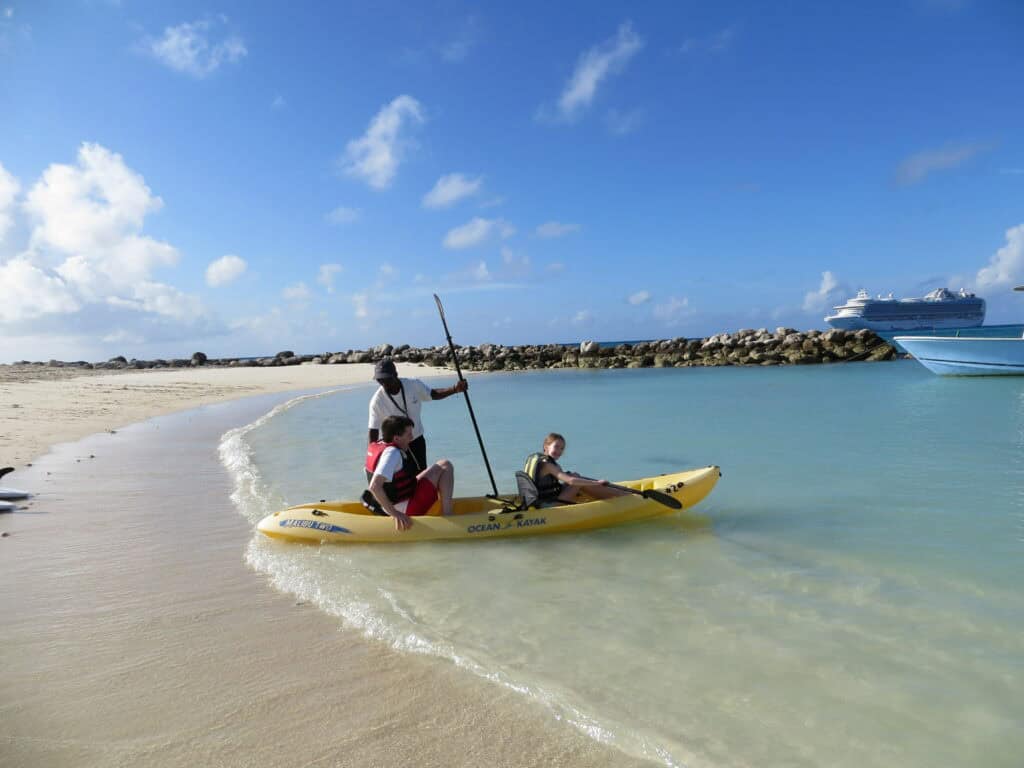 staff person holds yellow kayak while man and young girl in life vests get seated at edge of ocean with Crown Princess in background.