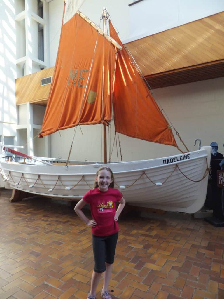 young girl in red shirt and dark long shorts poses in front of Madeleine boat at the Maritime Museum of the Atlantic.