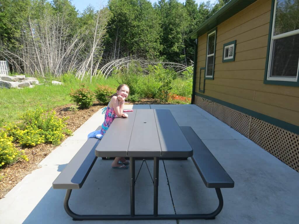 young girl sitting at picnic table on paved area alongside cabin.