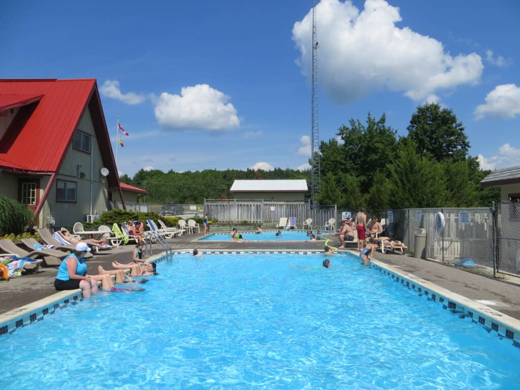 fenced in pool at Barrie KOA with building alongside, pool chairs, people sitting around pool and swimming.