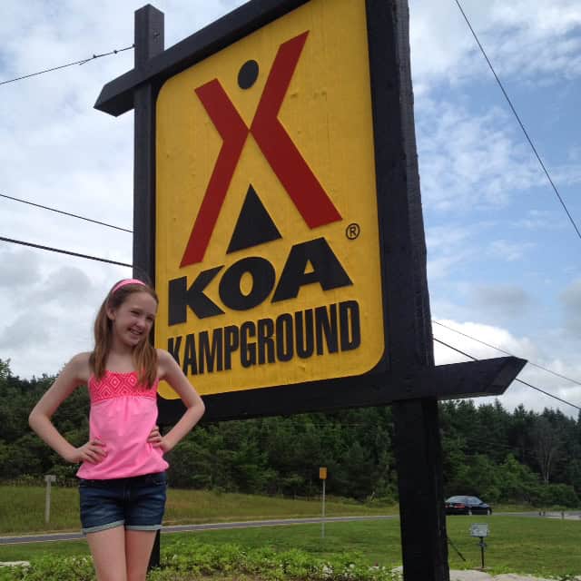 young girl in dark denim shorts and pink tank top poses with hands on hips by the yellow KOA Kampground sign - KOA Barrie.