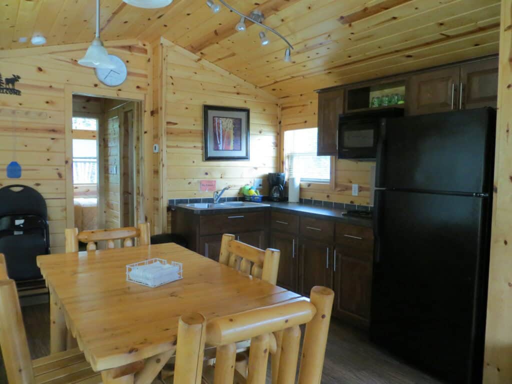 kitchen area in cabin - wall and ceiling wood panels with wood table and chairs, black fridge, dark cupboards and countertop.