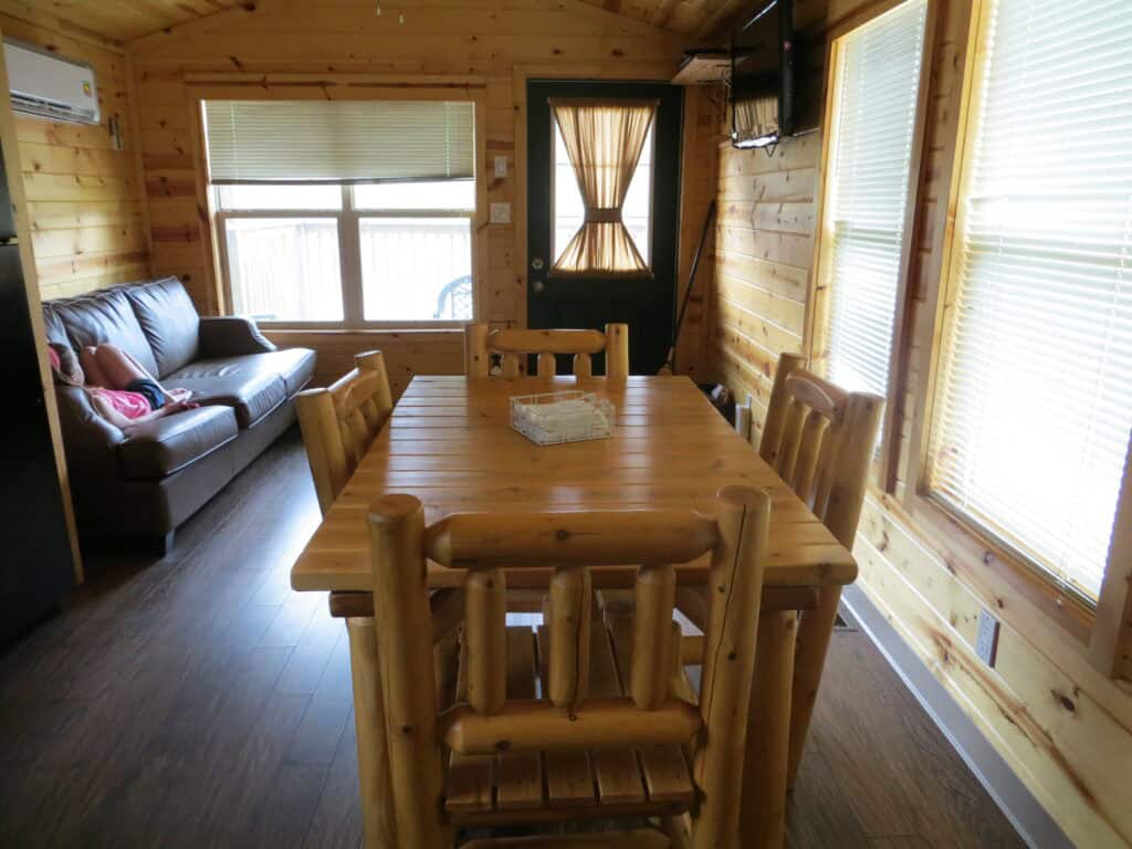 Barrie KOA - interior of Deluxe Cabin - wooden table with 4 wooden chairs, dark floors, wood walls, blinds on windows, dark couch.
