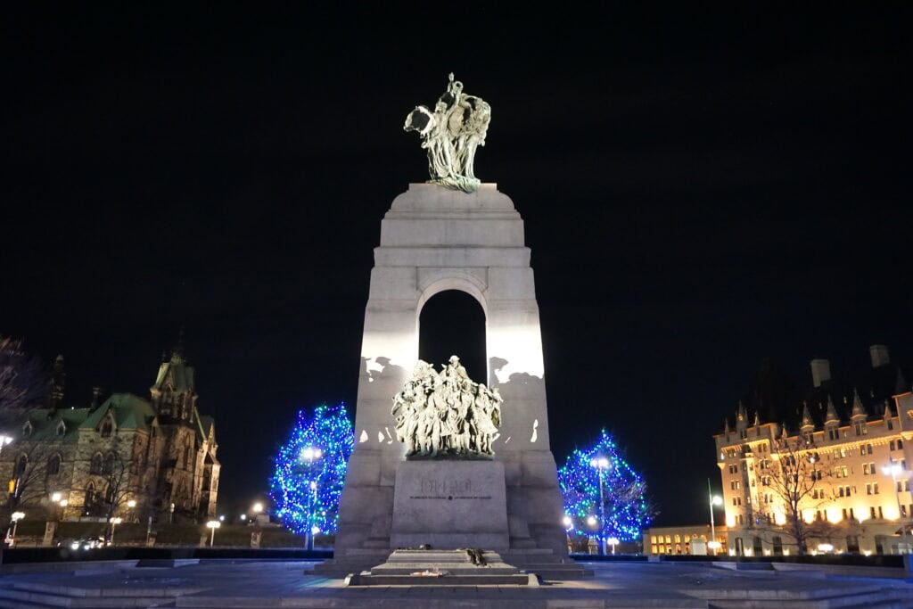 national war memorial in ottawa at night set against dark sky with two trees alongside with blue christmas lights and chateau laurier and parliament in background.