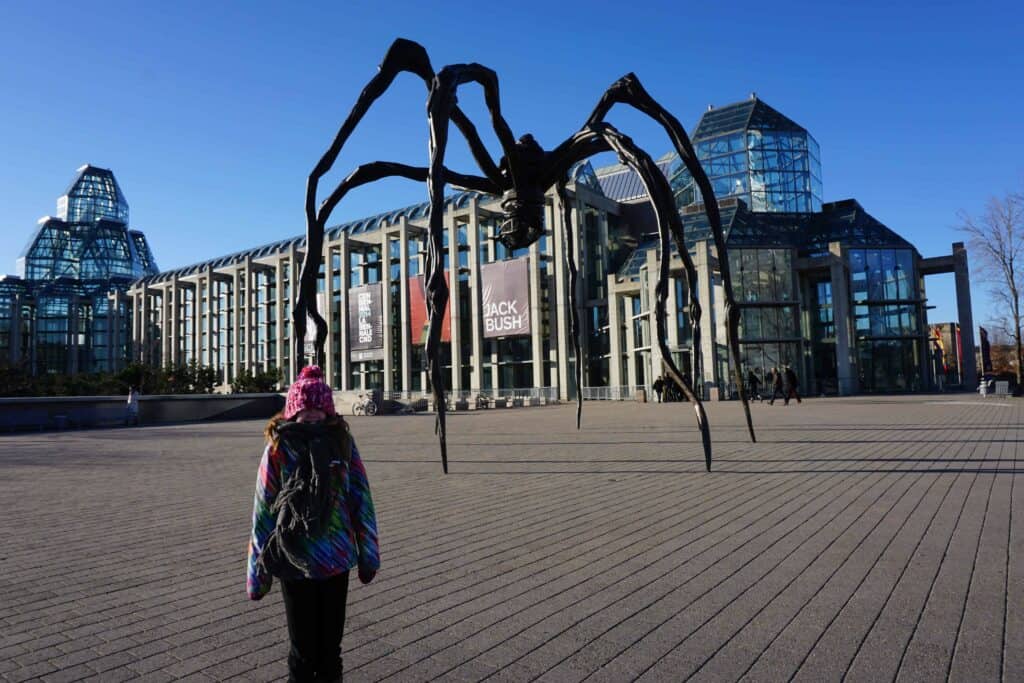 young girl bundled up in cold in front of national gallery and maman spider sculpture with bright blue sky.
