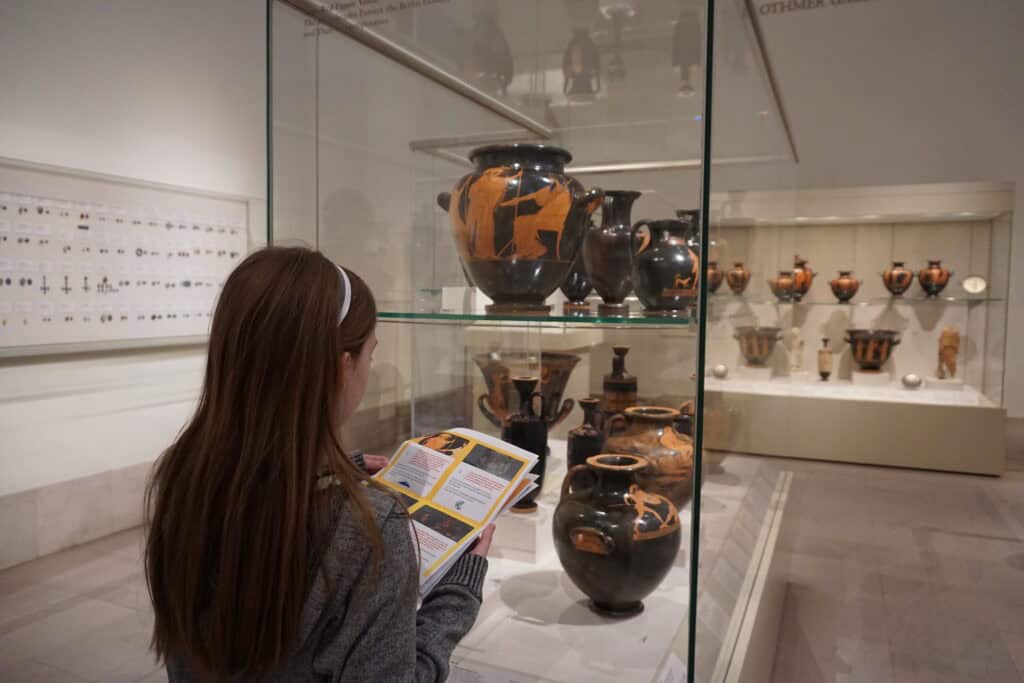young girl in grey sweater looks at Greek pottery while complete the Percy Jackson scavenger hunt at the Met in New York City.
