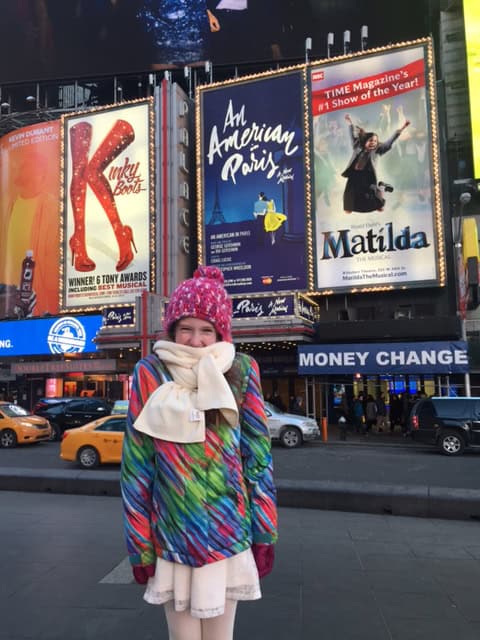 young girl in Times Square bundled up in colourful winter coat, dark pink hat and light pink skarf over pale pink skirt with Broadway posters in background.