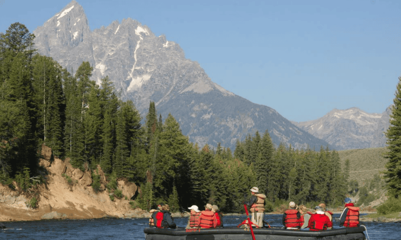 group of people wearing life vests most sitting but one standing in middle of river raft with mountains in background.