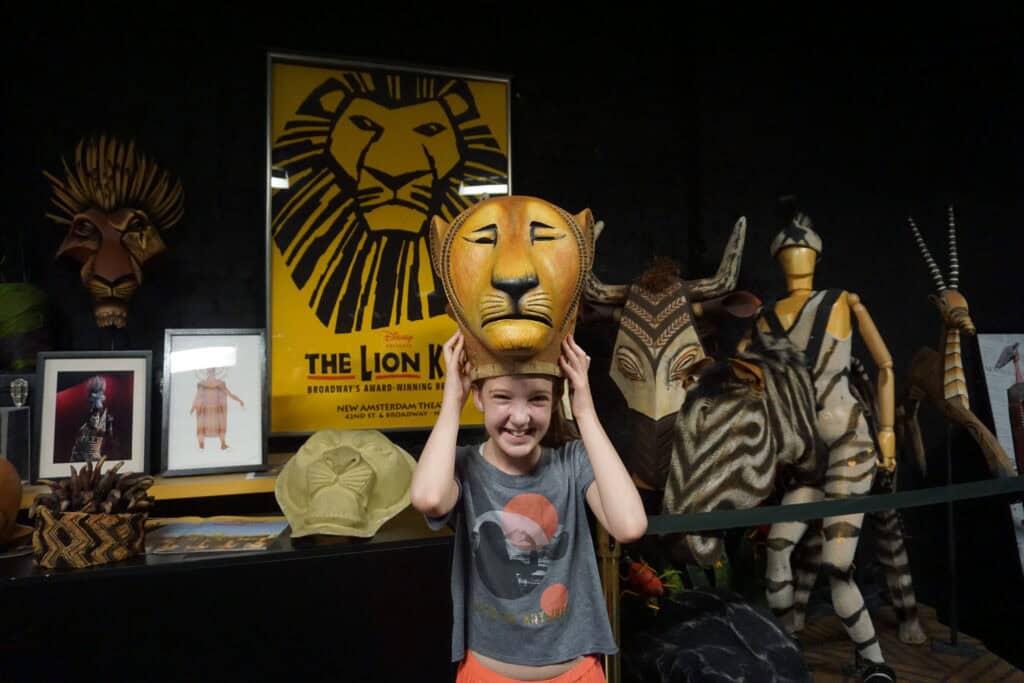 young girl trying on lion headdress in front of poster, props and zebra costume from The Lion King.