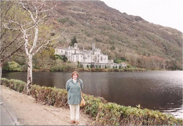 woman in beige pants and green coat standing beside water with Kylemore Abbey, Ireland in background.