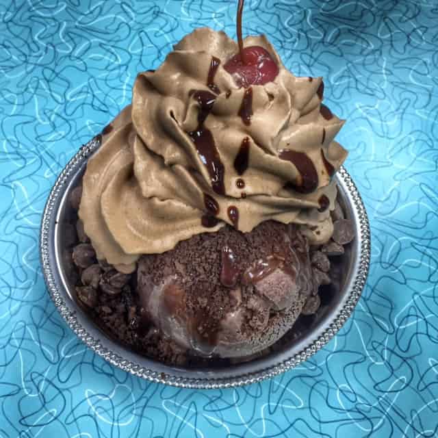 chocolate sundae with red cherry on top in silver metal dish on blue table with swirl design.