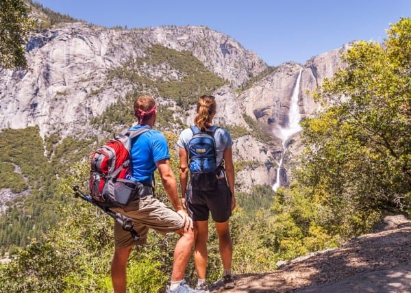 man and woman wearing shorts and t-shirts and carrying backpacks viewed from the back as they look at mountain range in yosemite national park.