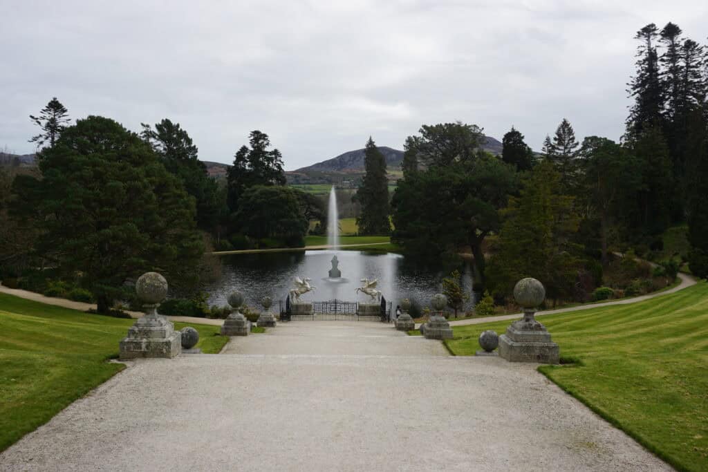 view down stepped walkway to lake with fountain in middle and winged horse at lake edge surrounded by trees and mountains in background on overcast day.