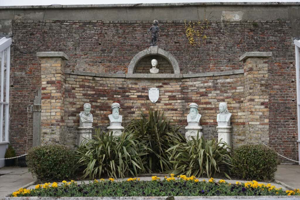 Julia's Memorial in Powerscourt Gardens - four busts of men in front of wall with bust of woman above - plants with yellow flowers.