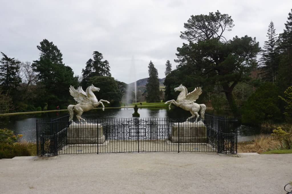 two wiwnged horses behind wrought iron gate alongside lake with fountain surrounded by trees - Powerscourt Gardens in Ireland.