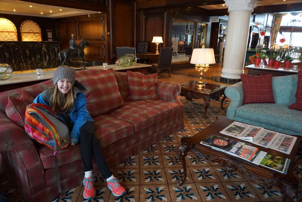 young girl in black pants, bright blue jacket, and grey wool beanie hat sits on red plaid sofa in lobby of the Powerscourt Hotel in Ireland.