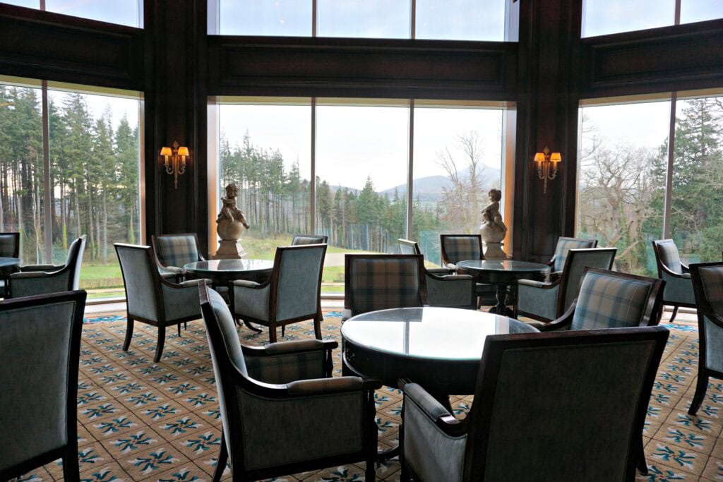 tables and chairs in lounge at the Powerscourt Hotel with large windows and view of trees and mountains in the distance.