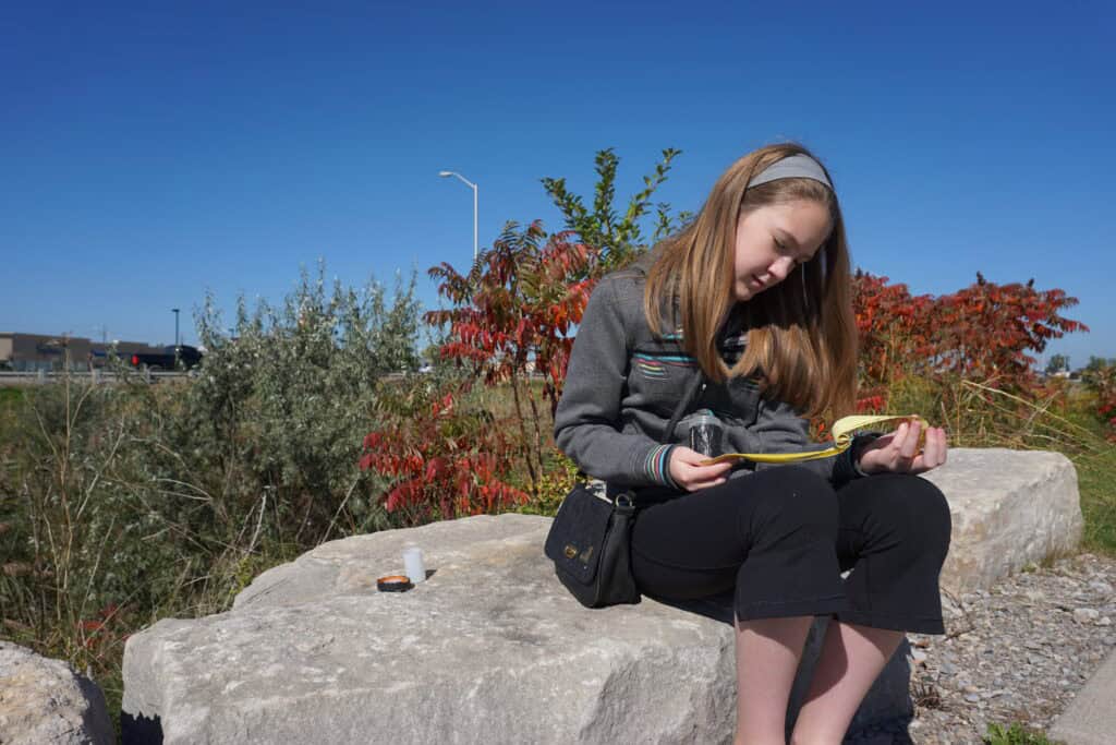 young girl with long hair and grey headband wearing black capri pants and grey hoodie sits on large rock looking at small book while holding geocache container in her hand.
