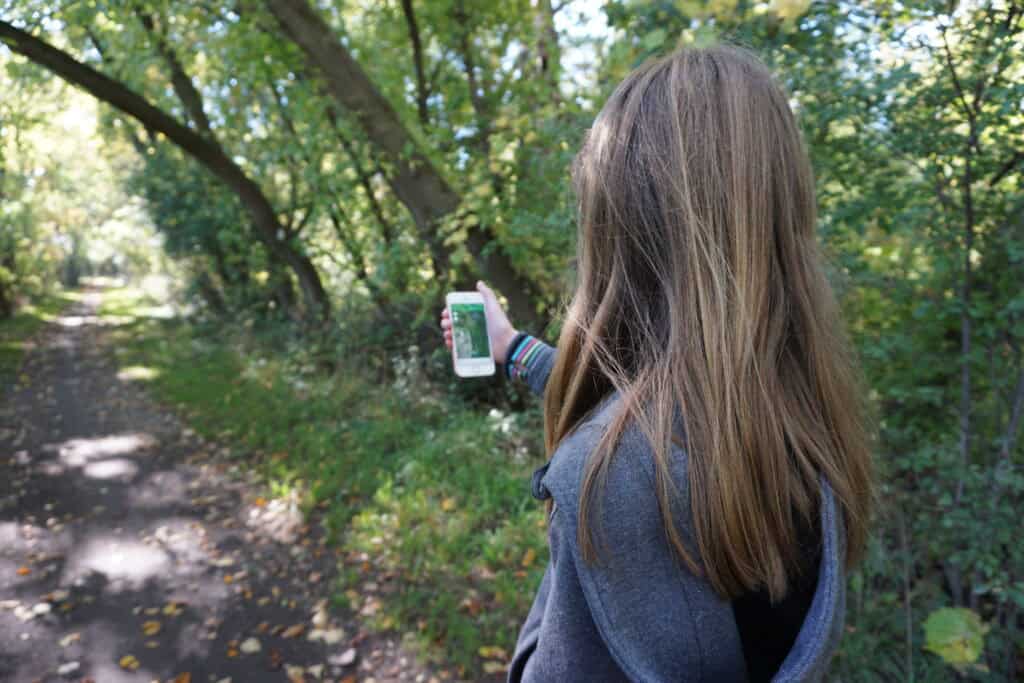 young girl on wooded trail holds iphone out in front of her with geocaching app on screen.