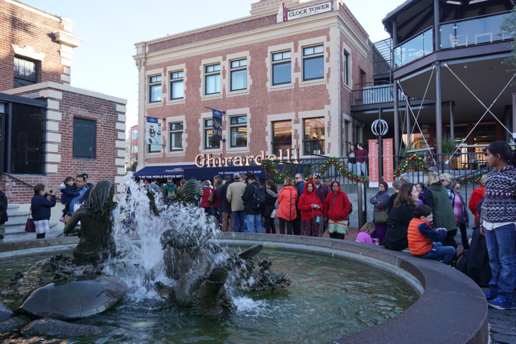 crowds of people around fountain in Ghirardelli Square with red brick Ghirardelli building in background - San Francisco.
