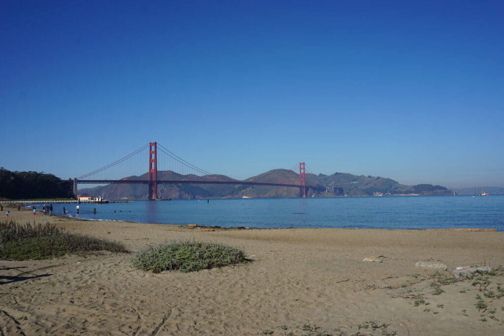 sandy beach of crissy field in foreground and view across bay to the golden gate bridge and mountains in background set against bright blue sky.