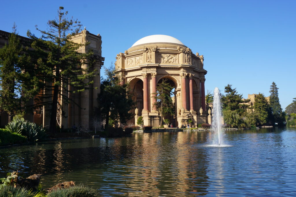 The Palace of Fine Arts in San Francisco - two toned reddish-brown rotunda with columns beside water with fountain set against bright blue cloudless sky.