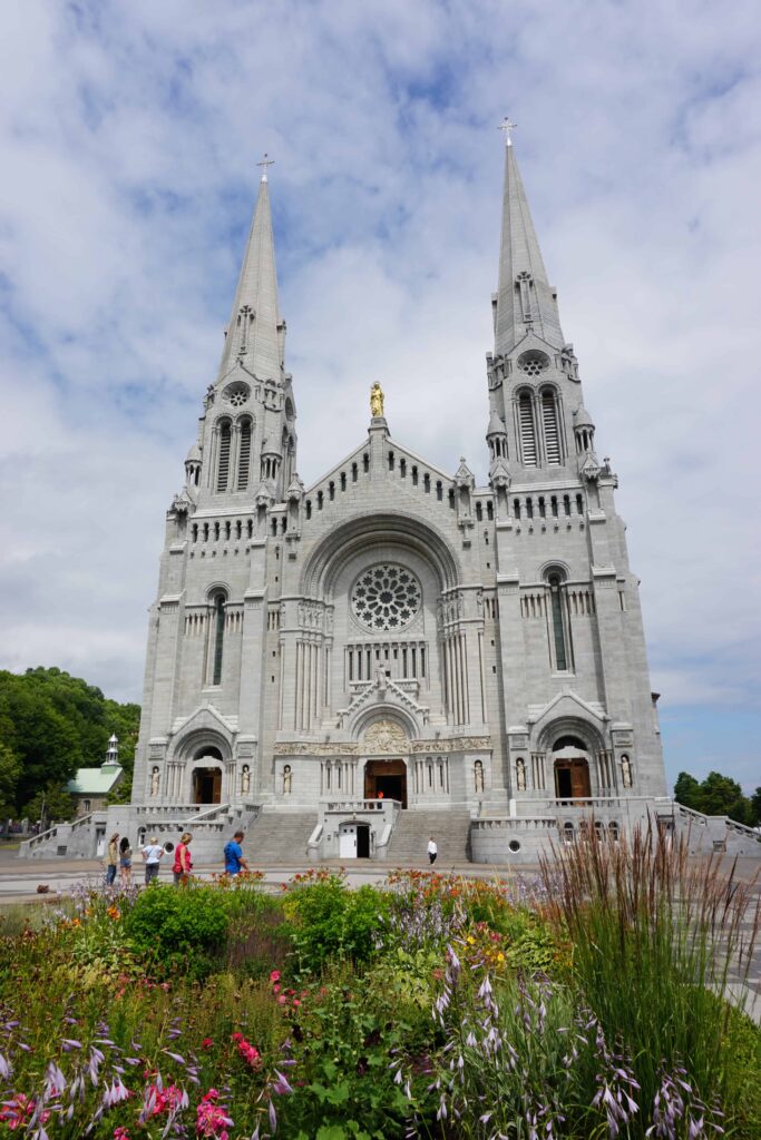 Flower gardens and a few people standing in front of the Basilica of Sainte-Anne-de-Beaupre in Quebec, Canada on a cloudy summer day.