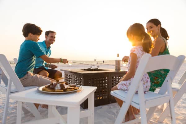 Family of mom, dad, boy and girl sit on white chairs on beach roasting marshmallows over a firepit.