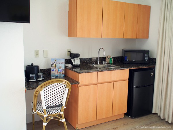 kitchenette at Sirata Beach Resort - light brown wood cuboards with bar sink, small black undercounter fridge, microwave, wall cupboards, small counter with coffee maker and chair.