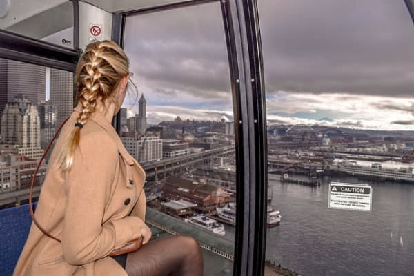 Blonde woman with hair in braid wearing camel coloured coat looks out window at Seattle waterfront from the Great Wheel.