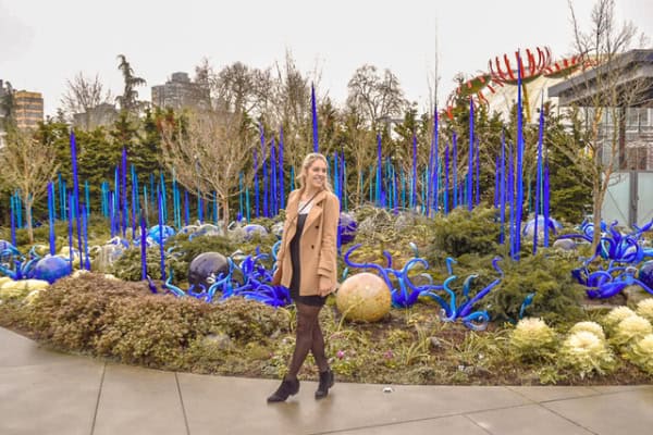 woman in black dress and camel coloured coat poses in front of bright blue glass sculptures in Chihuly Garden in Seattle.