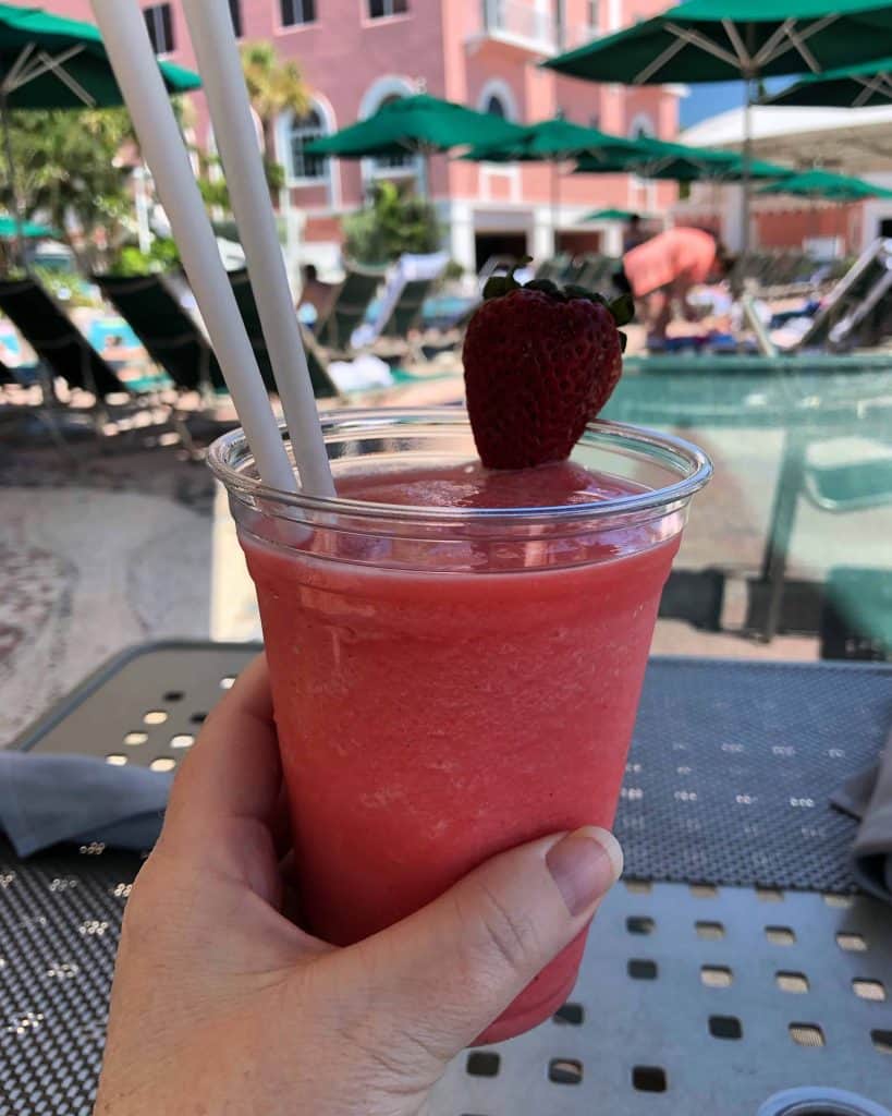 hand holding strawberry daiquiri with pink hotel and green umbrellas in background