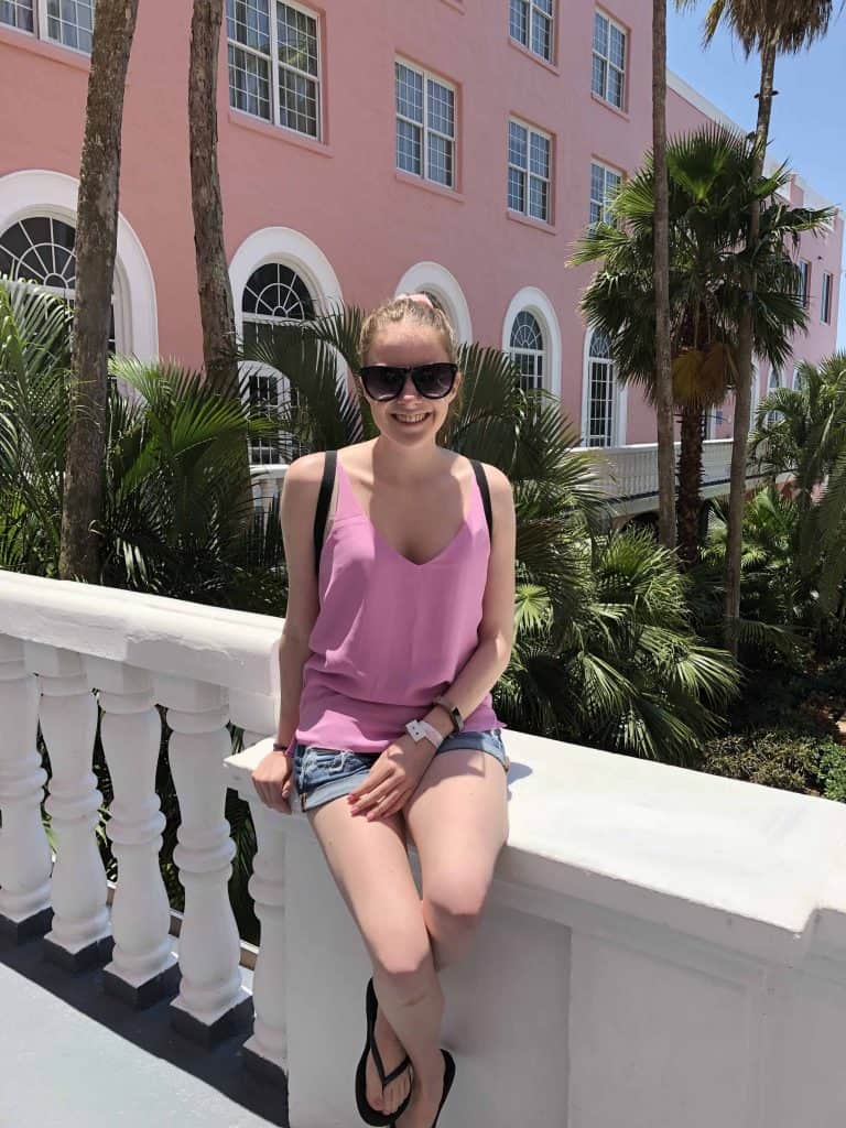 young woman in pink top sitting on white banister with pink hotel and palm trees in background