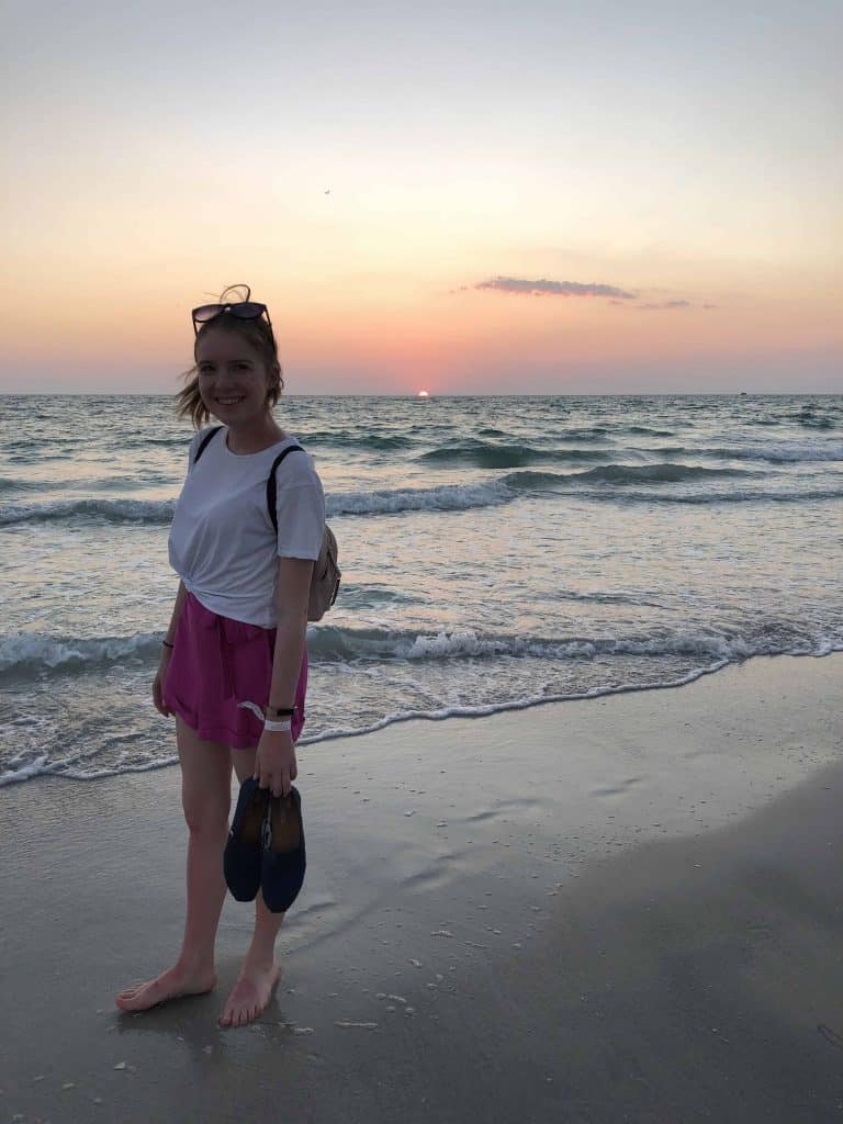 young woman in pink shorts and white shirt walking on beach at sunset