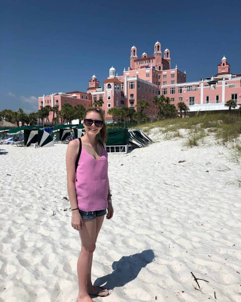 young woman in denim shorts and pink tank top on white sand beach with green beach umbrellas and pink hotel in background
