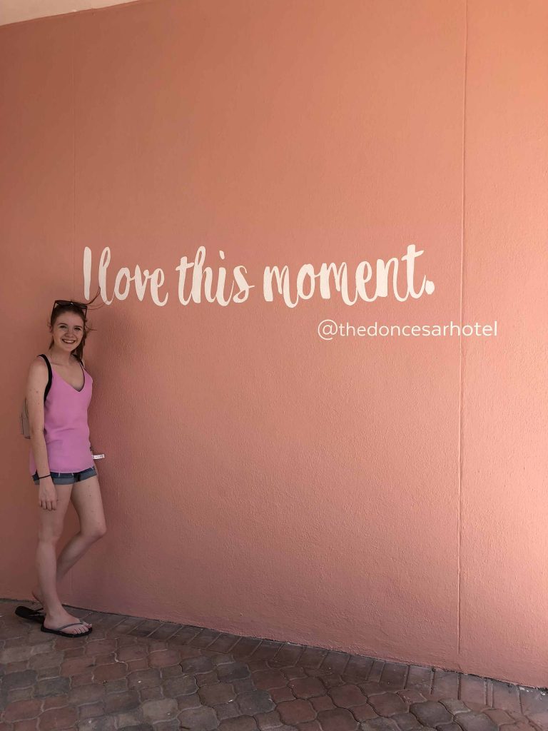 young woman pink top in front of pink wall with words I love this moment on wall