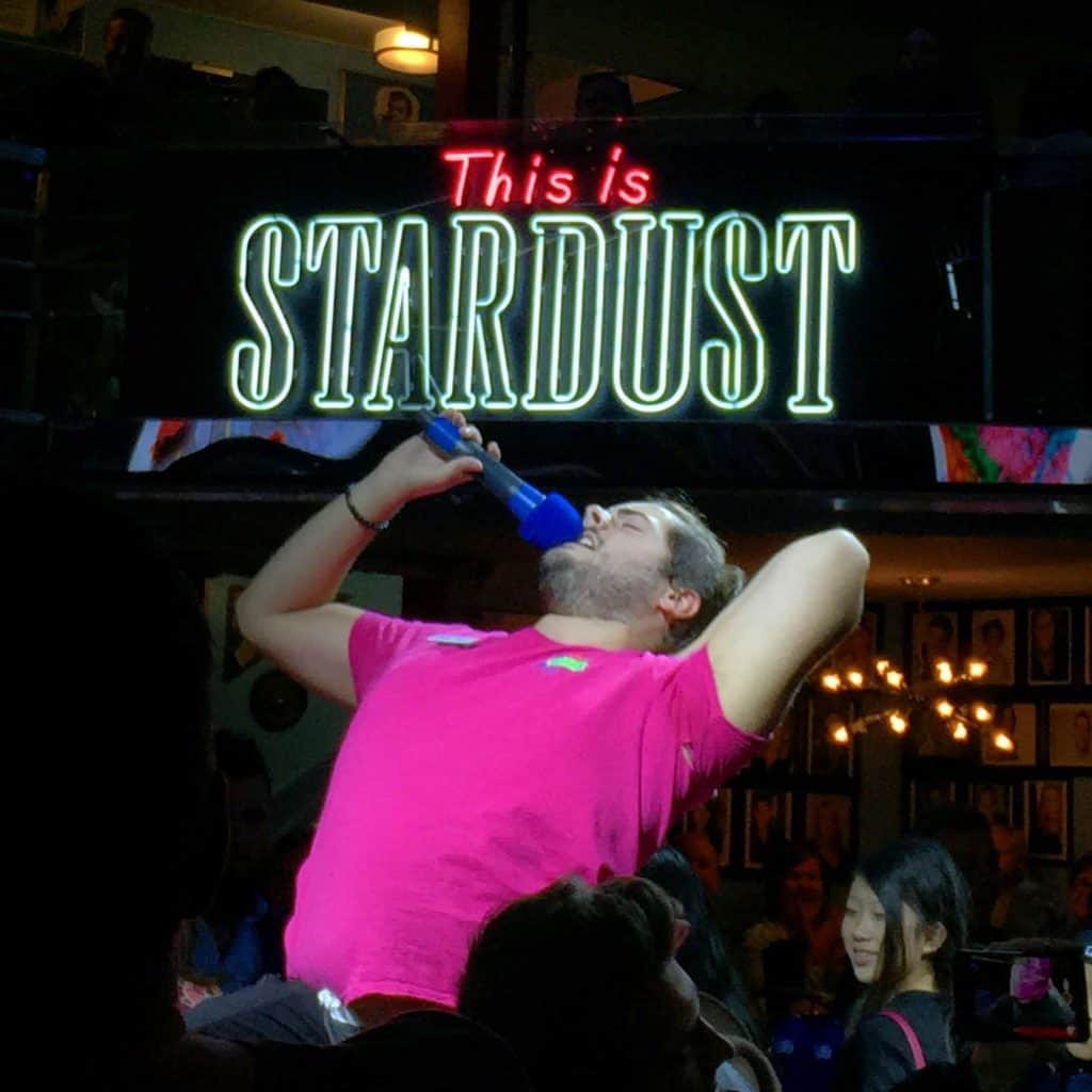 Man in pink shirt on table with microphone singing at Ellen's Stardust Diner in New York City.