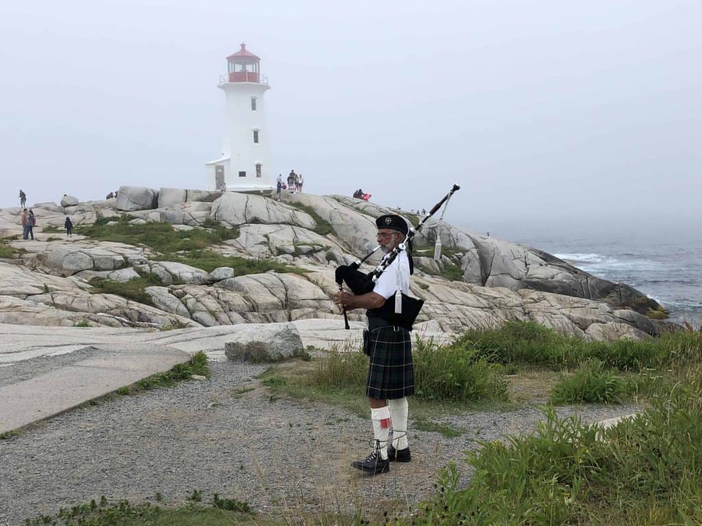 man in kilt playing bagpipes on rocks near red and white lighthouse