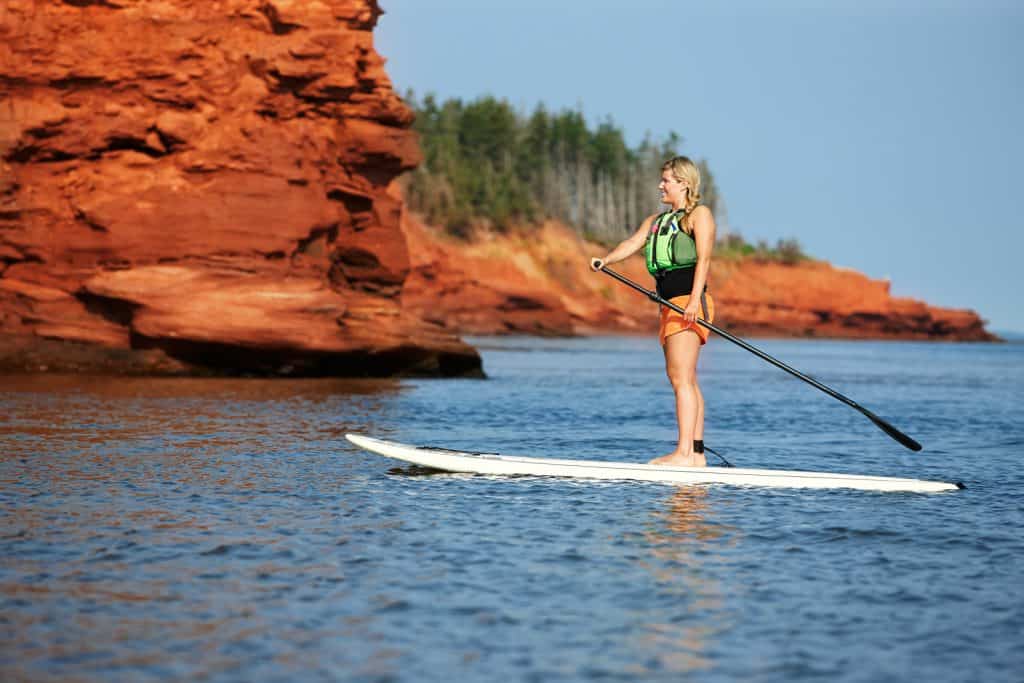 Young woman with paddle standing on white paddle board in ocean with red stone cliffs in background on Prince Edward Island.