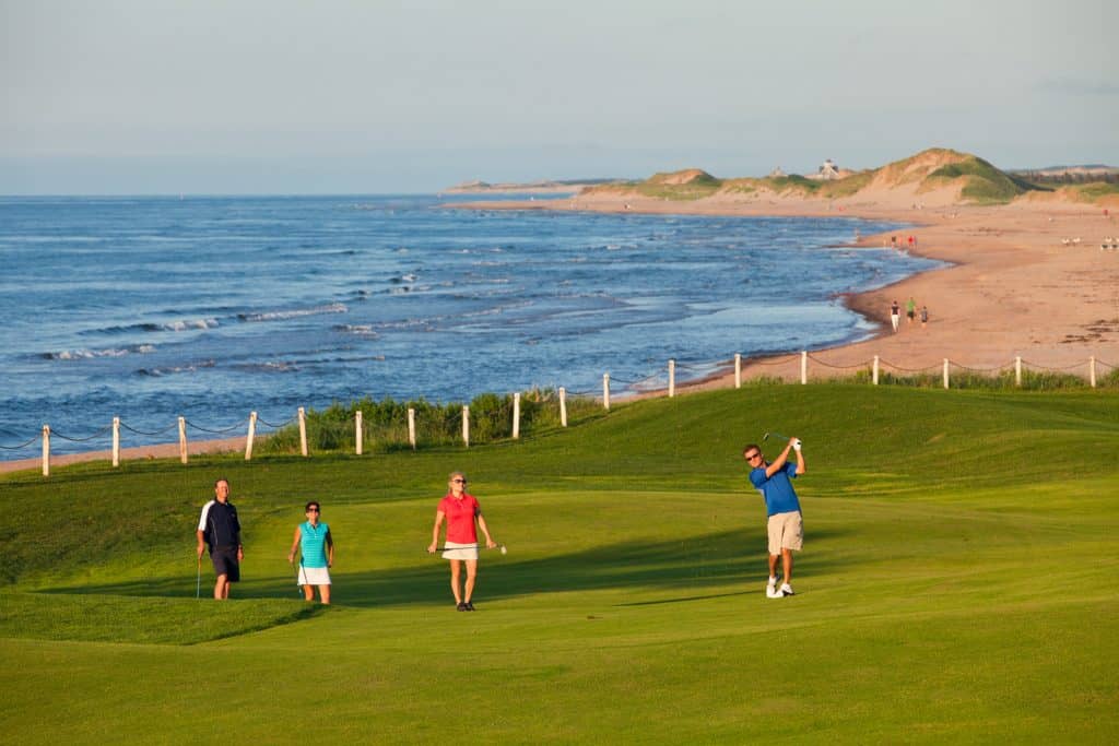 Golfer in mid-swing and three others watching on golf course in PEI with ocean and beach in background.