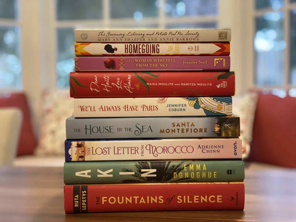Stack of books sitting on table with window seat and cushions in background.