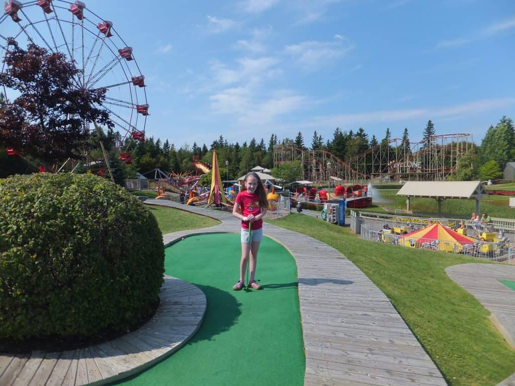 Young girl in light grey shorts and red t-shirt holding golf club standing on mini-putt course with rides in background at Sandspit amusement park in Cavendish, PEI.